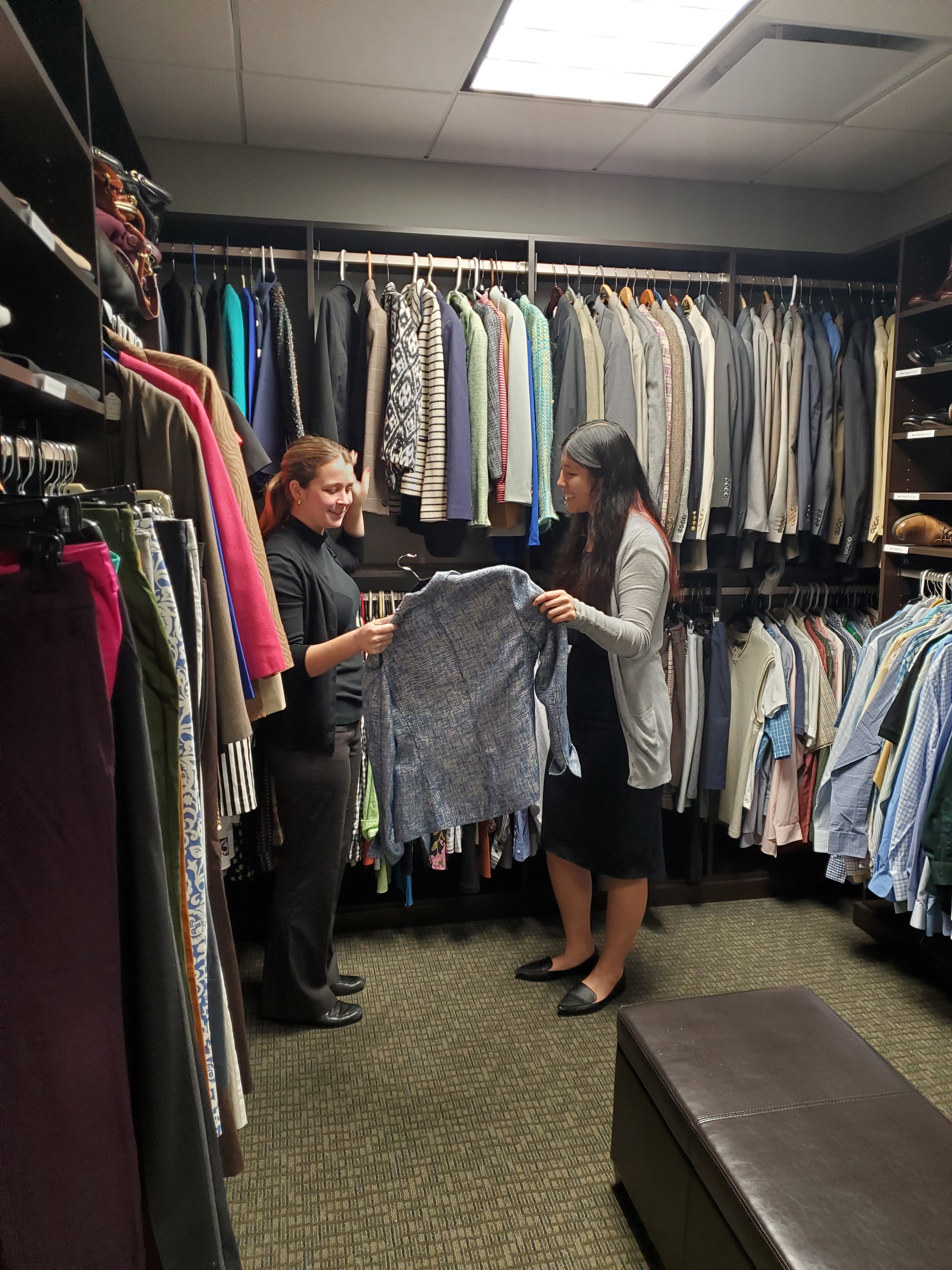 two women standing opposite each other inside of a room filled with suits.  One woman is handing a suit to another.